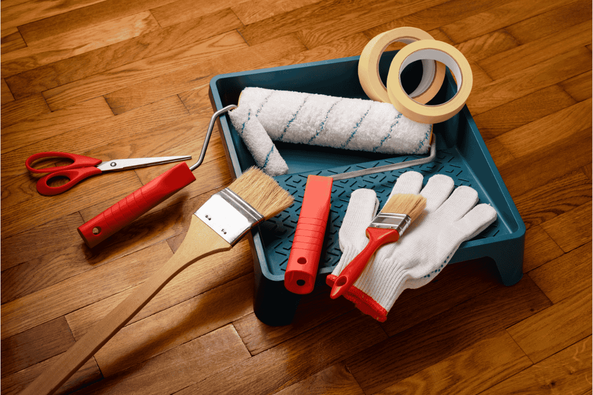 Painting tools including a paint roller, brushes, and gloves on a wooden floor.