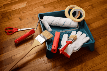 Painting tools including a paint roller, brushes, and gloves on a wooden floor.